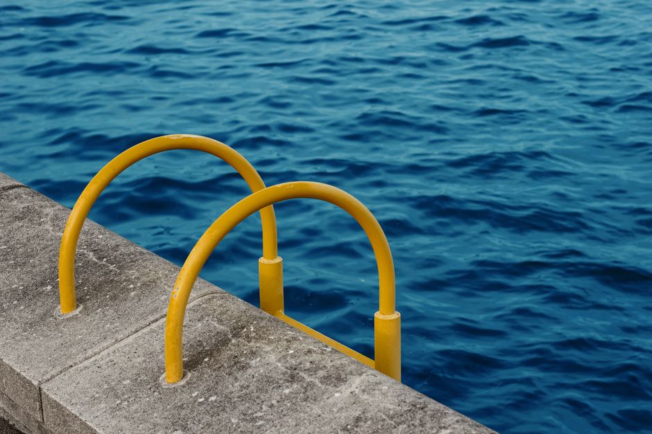 Close-up of a concrete dock with two yellow safety railings curved into semi-circular shapes, positioned at the edge overlooking the deep blue water. The concrete surface appears weathered but clean, 