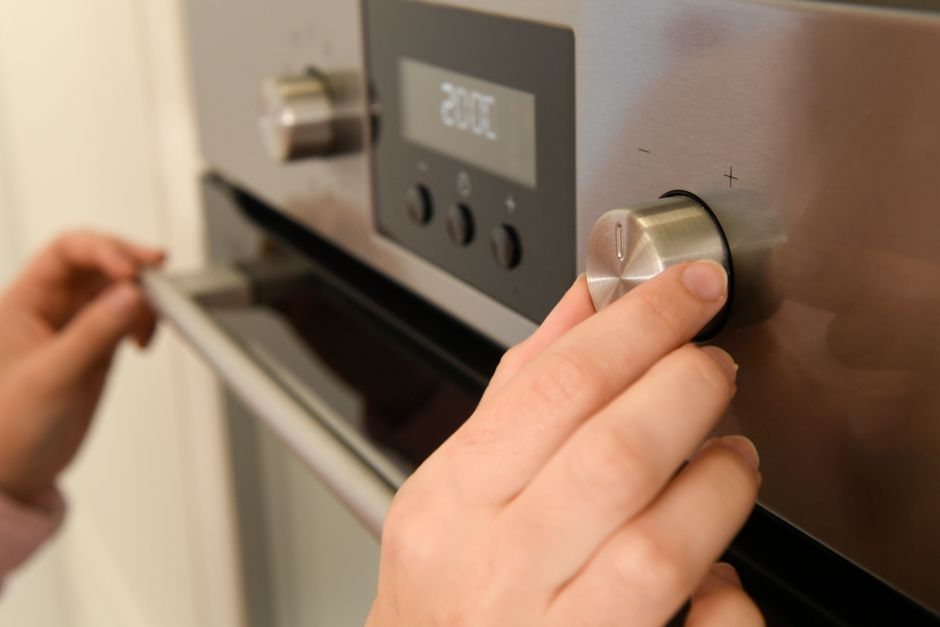 A woman wearing a white t-shirt, yellow and blue rubber cleaning gloves, and a black apron is seen in a modern kitchen, kneeling beside a black oven with the oven door open. She is holding a white spr