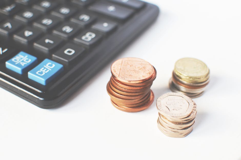 Close-up image showing a black calculator with light blue and white function keys, positioned on a white surface. Next to the calculator are three neatly stacked piles of coins—one pile of copper-colo